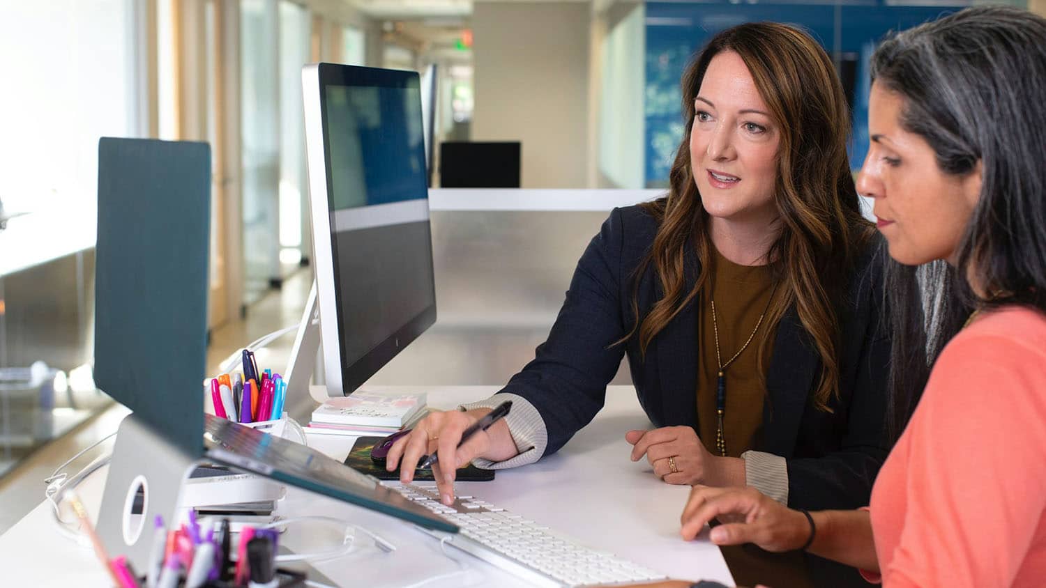 two women talk in an office while working on a computer